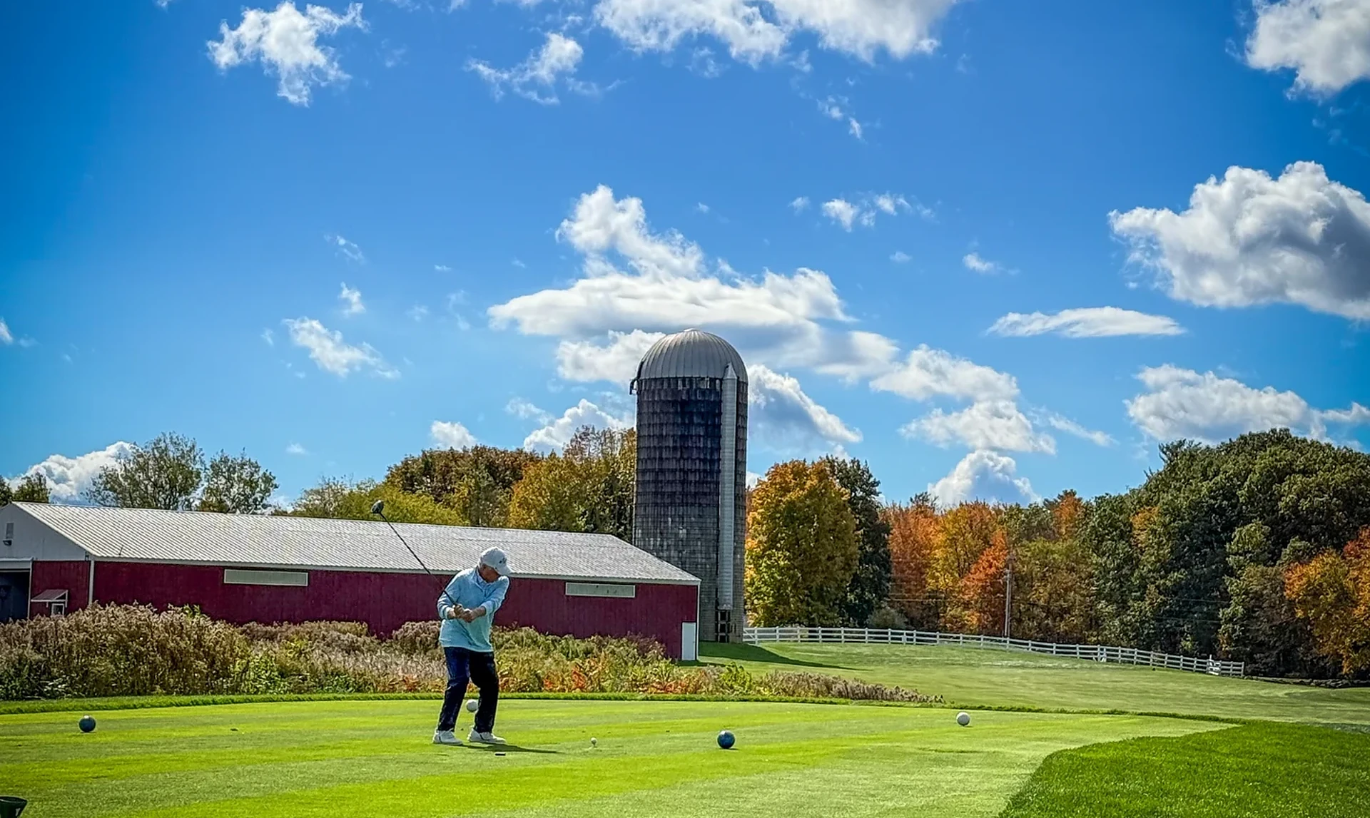 A golfer swings on the tee box in front of the red barn and silo at the 2026 AssMan Open